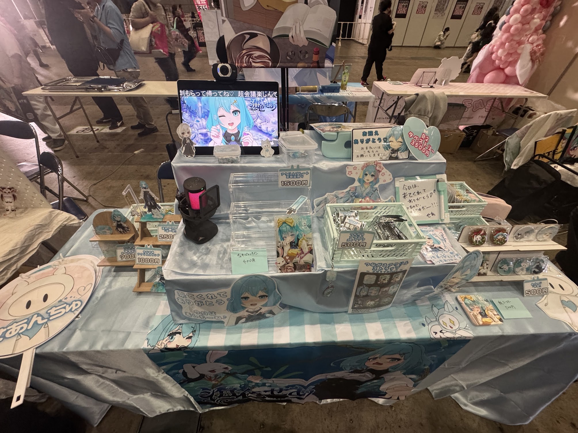 海色せあが映るモニターと、アクリルグッズや缶バッジなどが並んだイベントのブース。